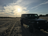 A black Jeep Wrangeler parked in the sandwithout the ocean visible, with the sun shining over the back of the car