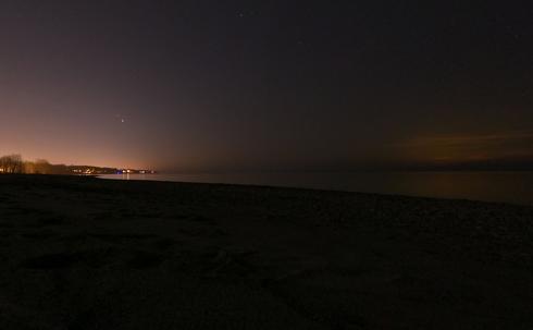 A picture taken parallel to the shoreline towards a town, glowing in the fog at night